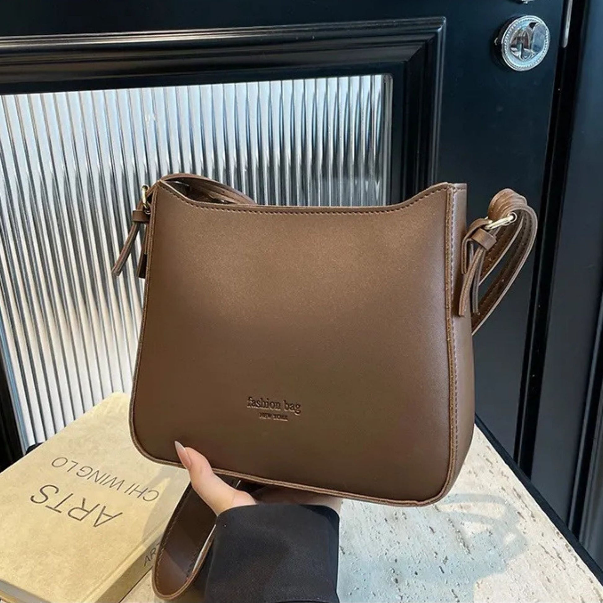 Brown shoulder bag held by a hand with a book in the background.