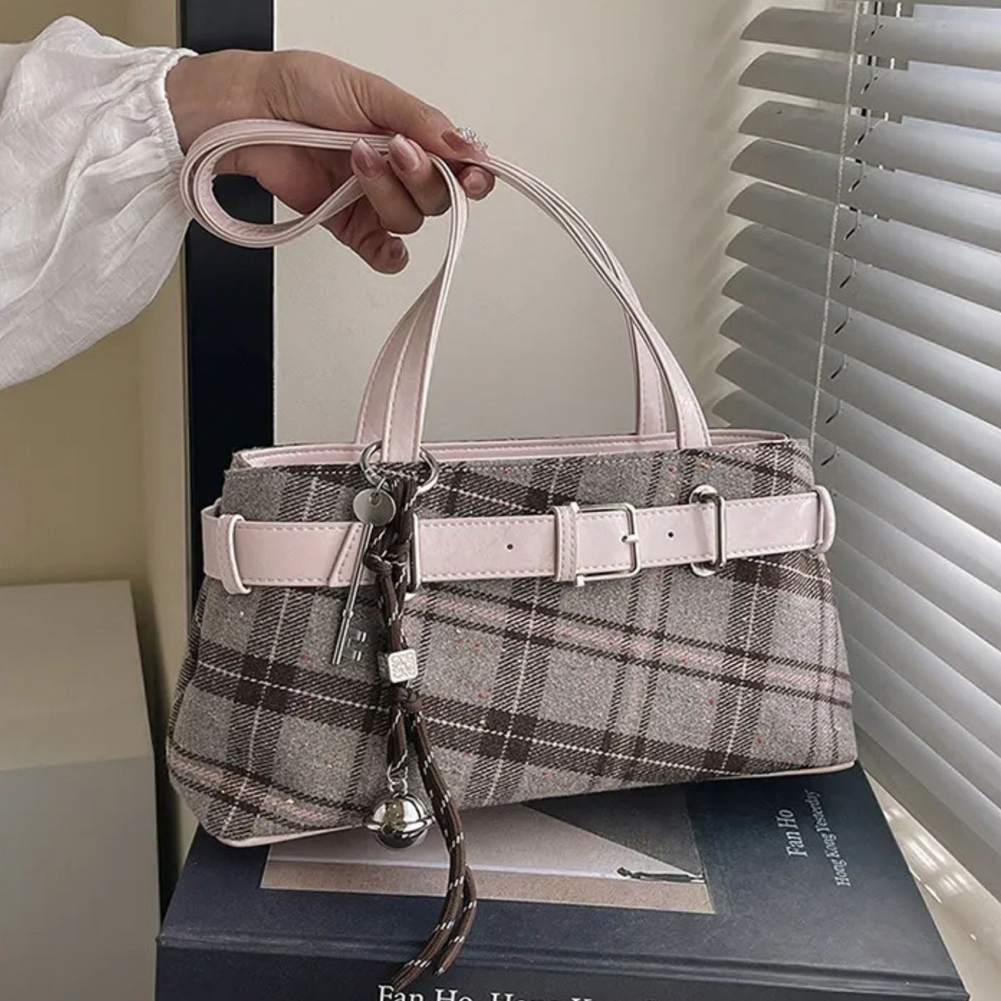Handbag with plaid pattern and pink handle held by a hand, placed on a book.