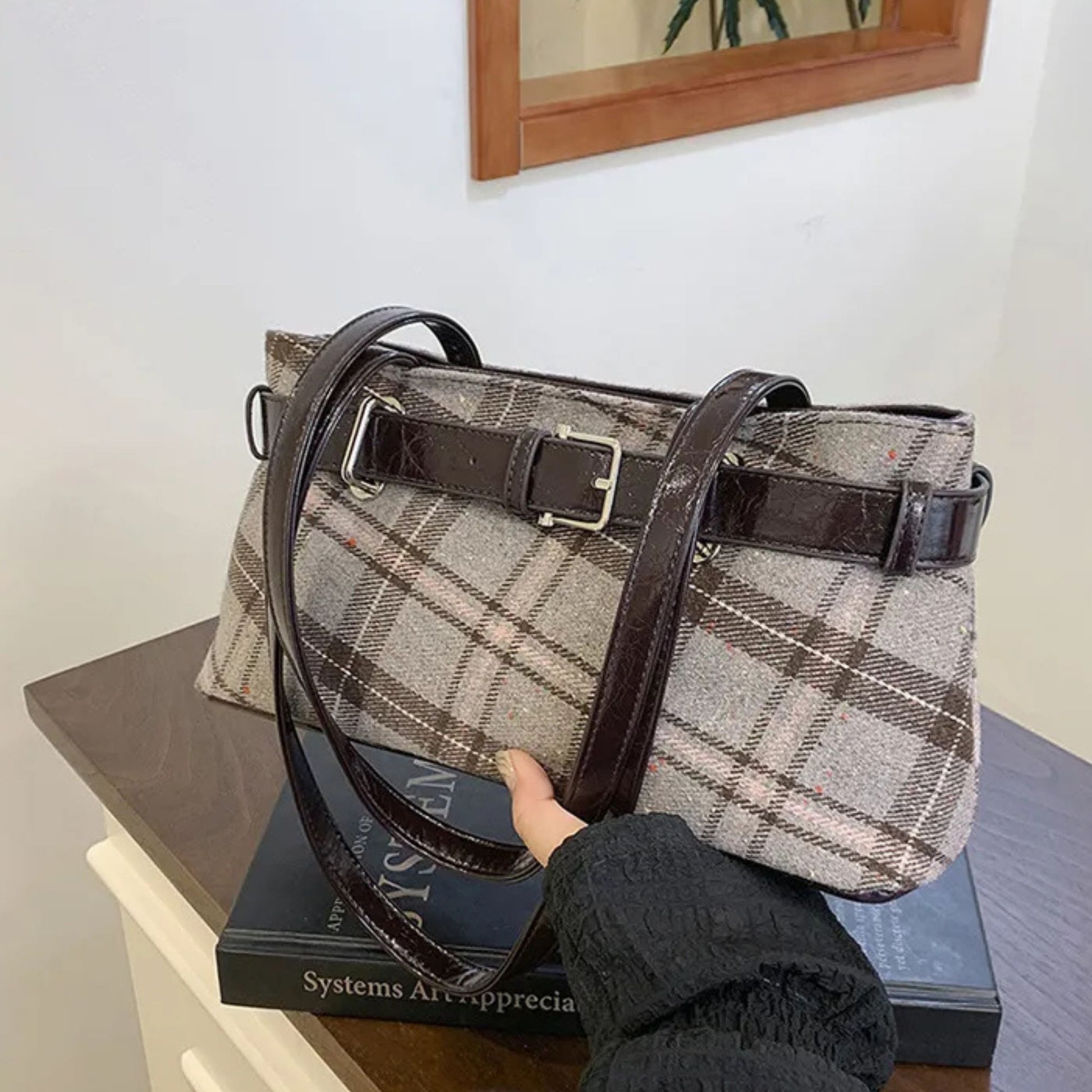 Plaid handbag with brown strap held by a person on a stack of books against a white wall.