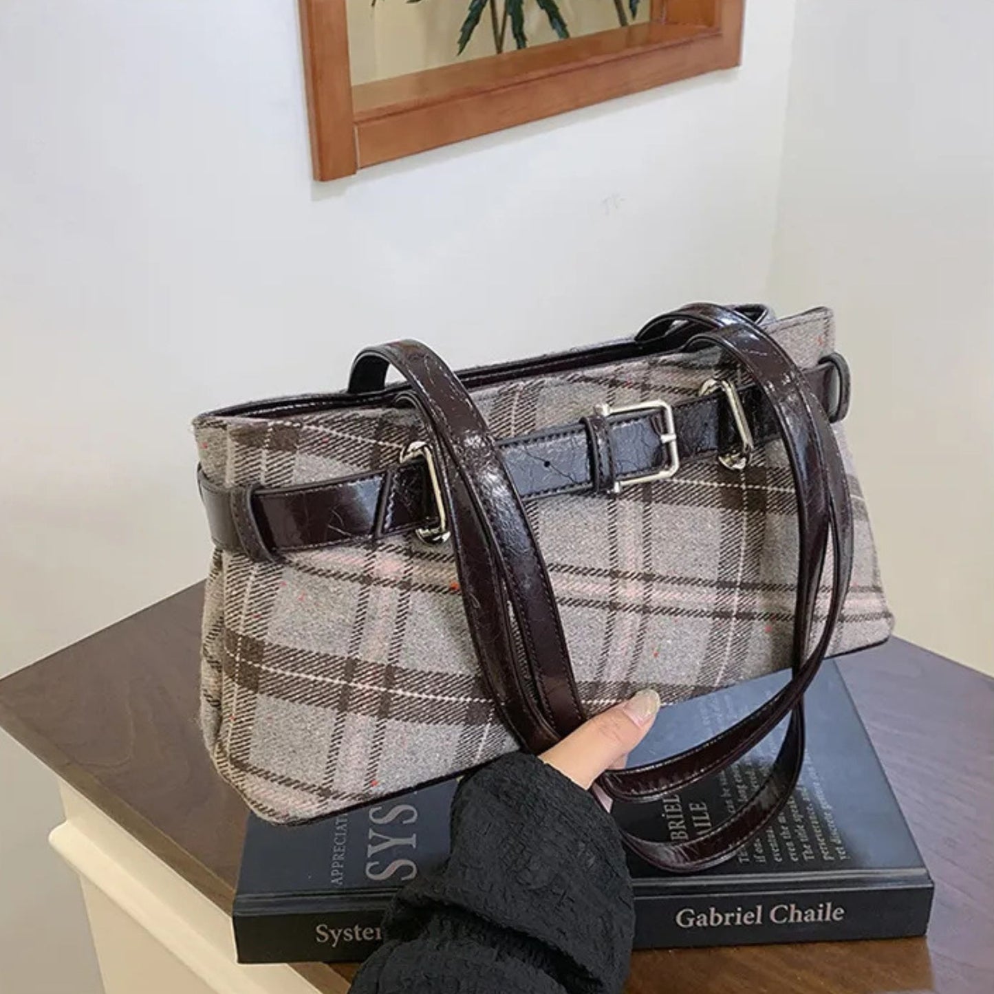 Plaid handbag with brown leather accents held over a book on a wooden surface.