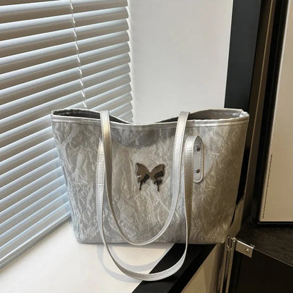 Silver textured totebag with a butterfly design on a surface near a window with blinds.