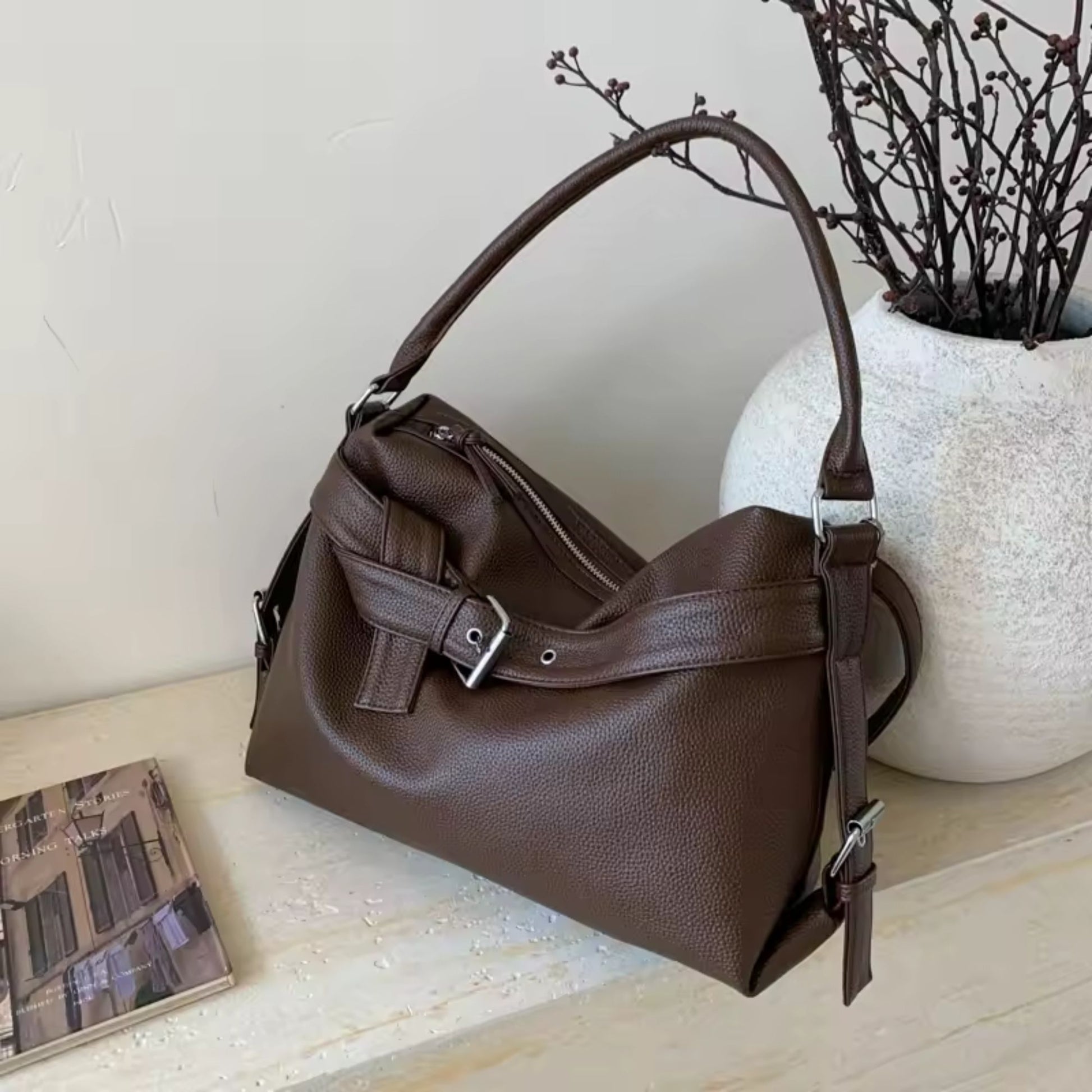 Brown leather large retro tote bag women on a light wooden surface with a white vase and dried branches in the background.