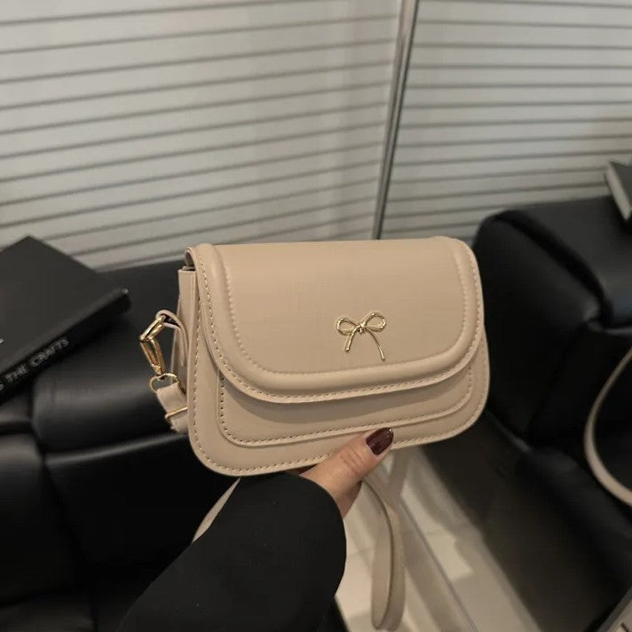 Beige shoulder bag with a bow detail held by a hand against a blurred indoor background
