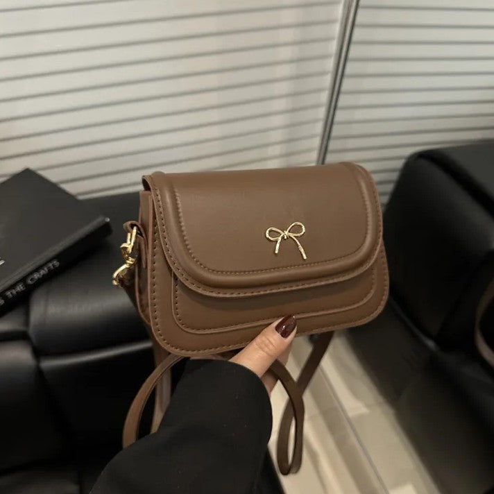 Brown shoulder bag with a gold bow held by a person against a neutral background